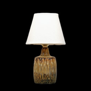 K&ouml;nnte beinhalten: Eine kleine Tischlampe mit wei&szlig;em Lampenschirm und Keramikfu&szlig;. Der Fu&szlig; hat eine braune und goldfarbene Glasur mit strukturierter Optik. Die Lampe ist f&uuml;r den Innenbereich konzipiert und bietet Umgebungslicht.