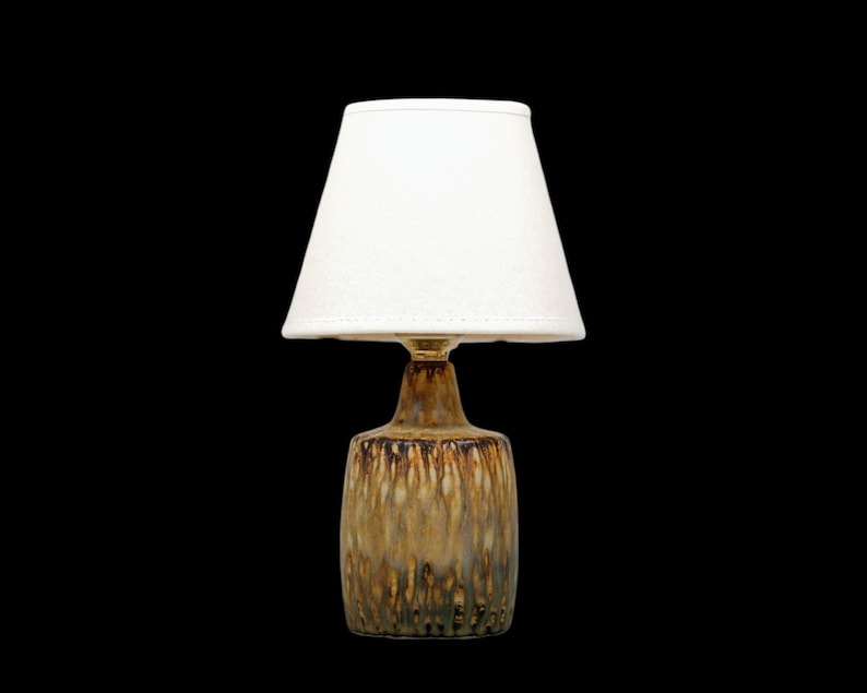 K&ouml;nnte beinhalten: Eine kleine Tischlampe mit wei&szlig;em Lampenschirm und Keramikfu&szlig;. Der Fu&szlig; hat eine braune und goldfarbene Glasur mit strukturierter Optik. Die Lampe ist f&uuml;r den Innenbereich konzipiert und bietet Umgebungslicht.