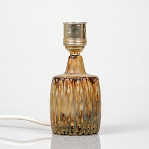 K&ouml;nnte beinhalten: Kleine Tischlampe aus Keramik mit brauner, beiger und gr&uuml;ner Glasur. Die Lampe hat einen zylindrischen Sockel und eine messingfarbene Fassung. Ein wei&szlig;es Stromkabel geht von der Basis aus.