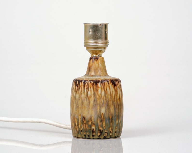 K&ouml;nnte beinhalten: Kleine Tischlampe aus Keramik mit brauner, beiger und gr&uuml;ner Glasur. Die Lampe hat einen zylindrischen Sockel und eine messingfarbene Fassung. Ein wei&szlig;es Stromkabel geht von der Basis aus.