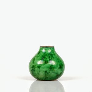 May include: A small, round, green vase with a speckled pattern. The vase has a narrow neck with a dark rim. The glossy finish reflects light, and the vase sits on a white surface.