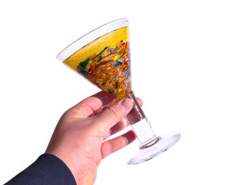 May include: A clear martini glass with a colorful abstract design inside. The glass has a wide, triangular bowl and a long stem. The interior of the glass features a yellow background with splashes of red, blue, and green.