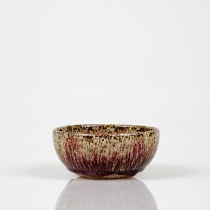 Puede incluir: Un pequeño cuenco de cerámica hecho a mano con un esmalte único. El borde es marrón claro moteado, que pasa a un borgoña profundo con vetas verticales. El interior es de un borgoña más oscuro. El cuenco es probablemente para servir comida.