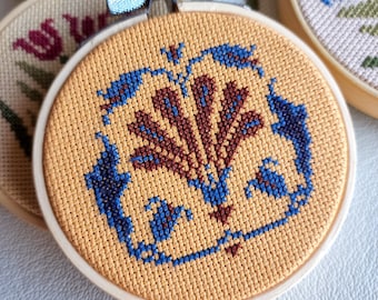 May include: A yellow embroidery hoop with a cross-stitch design of a stylized brown and blue flower. The flower is surrounded by a blue border.