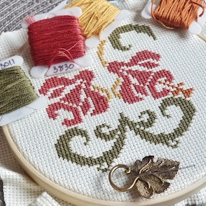 Puede incluir: Primer plano de un aro de bordado de punto de cruz con la palabra "Gather" bordada en hilo rojo, verde y amarillo. El aro está sobre una superficie blanca con carretes de hilo de bordar en rojo, amarillo y verde.