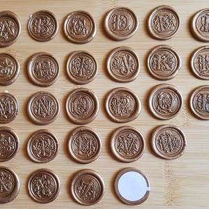 Puede incluir: Un conjunto de 36 sellos de cera dorada con diferentes iniciales de letras. Los sellos están dispuestos en una cuadrícula sobre una superficie de madera.