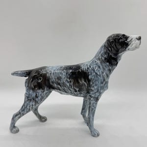 Op de afbeelding: Een keramieken beeldje van een hond in staande houding. De hond heeft een gespikkeld patroon van zwart en wit, met een witte vlek op zijn snuit. Het beeldje is gedetailleerd en lijkt een jachthondenras te zijn.