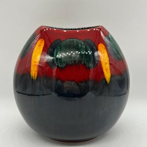 Könnte beinhalten: Eine dekorative Keramikvase mit runder Form. Die Vase hat eine glänzende Oberfläche mit einem Farbschema aus Rot, Schwarz, Grün und Gelb. Die Oberseite ist rot, geht am Boden in Schwarz über, mit farbigen Akzenten.