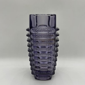 Pode incluir: Um vaso de vidro roxo translúcido com um design geométrico distinto, apresentando faixas horizontais caneladas e uma abertura quadrada. O corpo do vaso exibe um padrão repetitivo de cristas arredondadas em relevo nas laterais.