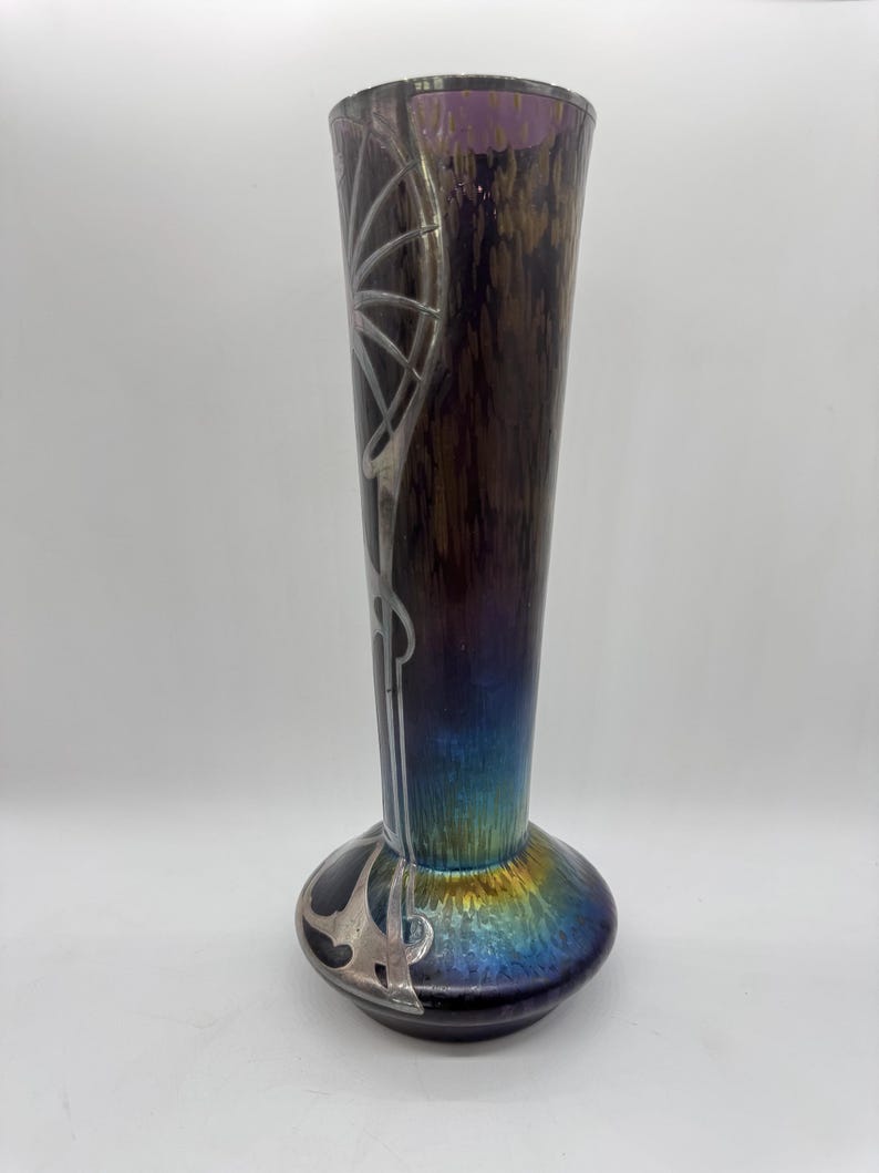 Pu&ograve; includere: Un vaso alto in vetro artistico iridescente con base bulbosa e collo sottile. Il vaso presenta un design Art Nouveau argentato e una sfumatura di colore dal viola al blu all'oro. Il vaso &egrave; alto circa 30 cm.