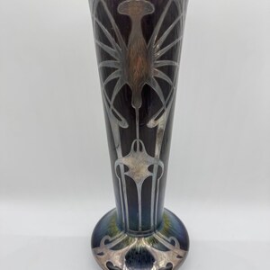 Pu&ograve; includere: Un vaso alto in stile Art Nouveau con la parte superiore svasata e una base arrotondata. Il vaso presenta uno sfondo scuro e iridescente con disegni argentati, stilizzati, floreali e simili a insetti. La base ha un bordo decorativo argentato.