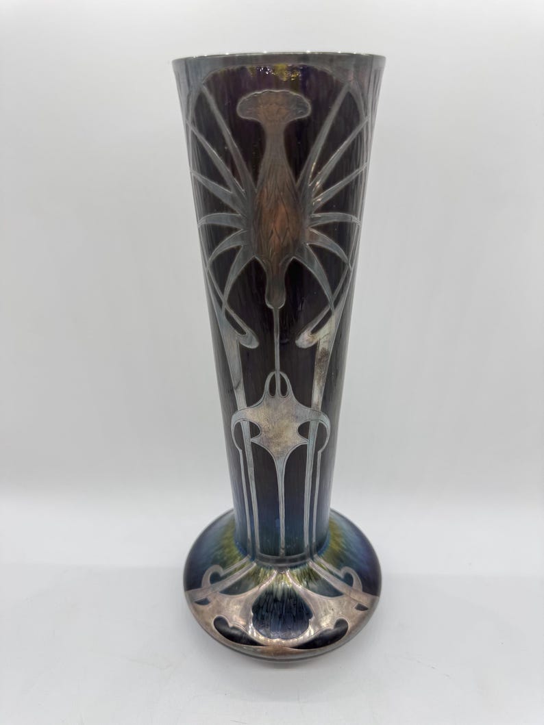Pu&ograve; includere: Un vaso alto in stile Art Nouveau con la parte superiore svasata e una base arrotondata. Il vaso presenta uno sfondo scuro e iridescente con disegni argentati, stilizzati, floreali e simili a insetti. La base ha un bordo decorativo argentato.