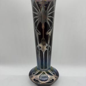 Pu&ograve; includere: Un vaso alto in stile Art Nouveau con finitura scura e iridescente. Il vaso presenta disegni floreali e geometrici stilizzati color argento. La base &egrave; arrotondata e il collo &egrave; lungo e sottile. Il vaso &egrave; alto circa 30 cm.