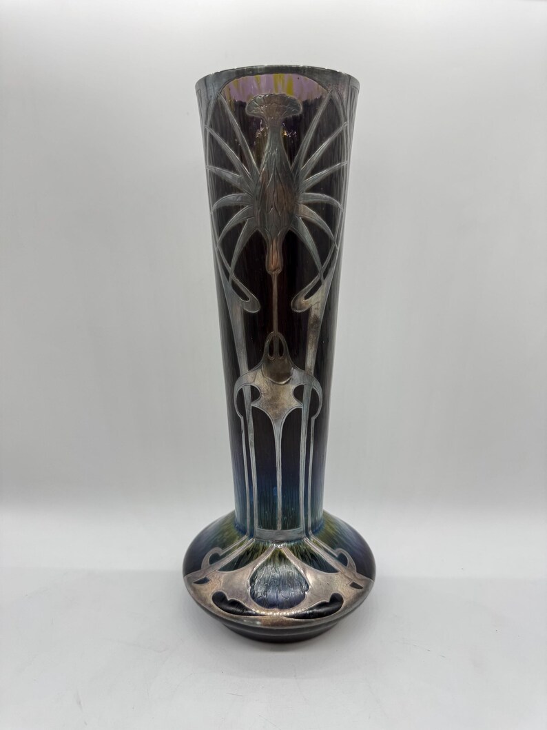 Pu&ograve; includere: Un vaso alto in stile Art Nouveau con finitura scura e iridescente. Il vaso presenta disegni floreali e geometrici stilizzati color argento. La base &egrave; arrotondata e il collo &egrave; lungo e sottile. Il vaso &egrave; alto circa 30 cm.