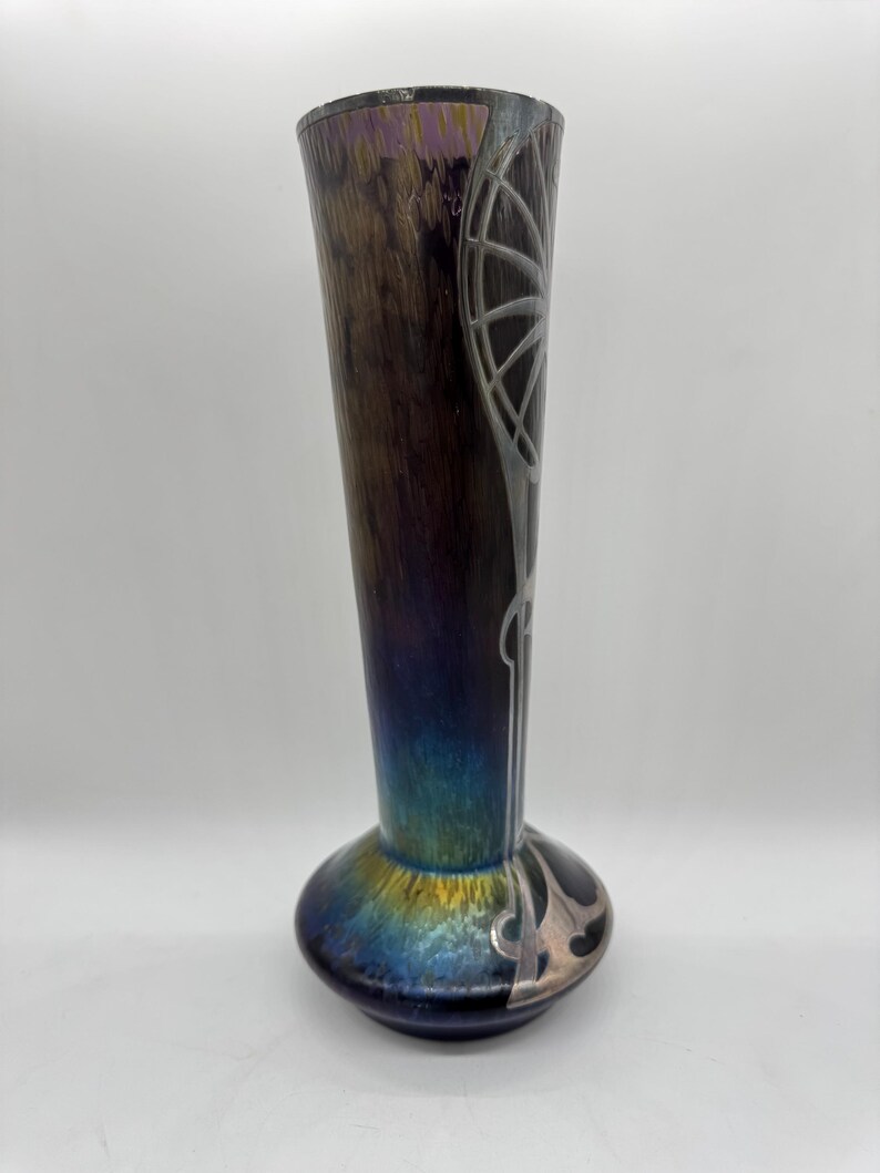 Pu&ograve; includere: Un vaso alto in vetro artistico iridescente con la parte superiore svasata e una base bulbosa. Il vaso presenta una combinazione di colori marrone scuro e viola con abbellimenti argentati in stile Art Nouveau. Il vaso &egrave; alto circa 30 cm.