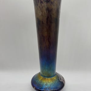 Pu&ograve; includere: Un vaso alto in vetro iridescente con la parte superiore svasata e una base bulbosa. Il vaso ha una transizione di colore dal blu scuro alla base al marrone e viola nella parte superiore. La base presenta un disegno decorativo. Un pezzo decorativo per la casa, alto circa 30 cm.