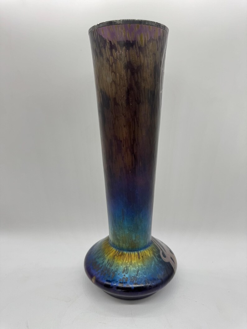 Pu&ograve; includere: Un vaso alto in vetro iridescente con la parte superiore svasata e una base bulbosa. Il vaso ha una transizione di colore dal blu scuro alla base al marrone e viola nella parte superiore. La base presenta un disegno decorativo. Un pezzo decorativo per la casa, alto circa 30 cm.