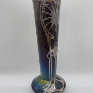 Pu&ograve; includere: Un vaso alto in vetro artistico iridescente con base bulbosa e parte superiore svasata. Il vaso presenta un corpo marrone scuro e viola con disegni floreali e geometrici argentati in stile Art Nouveau. La base passa al blu e all'oro.