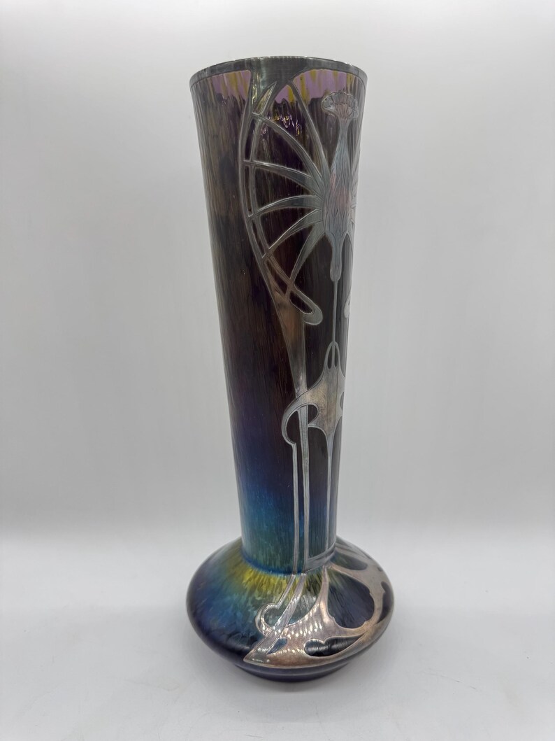 Pu&ograve; includere: Un vaso alto in vetro artistico iridescente con base bulbosa e parte superiore svasata. Il vaso presenta un corpo marrone scuro e viola con disegni floreali e geometrici argentati in stile Art Nouveau. La base passa al blu e all'oro.