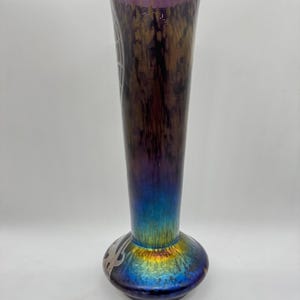 Pu&ograve; includere: Un vaso alto in vetro iridescente con la parte superiore svasata e una base bulbosa. Il vaso ha una transizione di colore dal viola scuro nella parte superiore al blu vibrante e oro nella base. Presenta disegni argentati incisi.