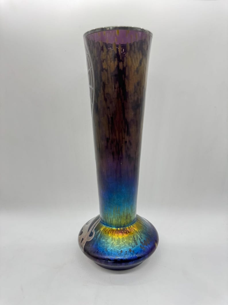 Pu&ograve; includere: Un vaso alto in vetro iridescente con la parte superiore svasata e una base bulbosa. Il vaso ha una transizione di colore dal viola scuro nella parte superiore al blu vibrante e oro nella base. Presenta disegni argentati incisi.