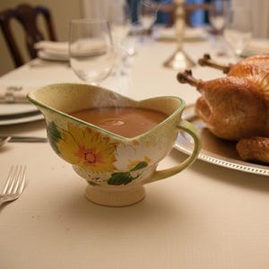 May include: A roasted turkey on a silver platter and a gravy boat filled with gravy are on a table set for a meal. The gravy boat has a floral design with yellow and white flowers. Silverware and plates are also on the table.