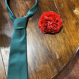 May include: A dark green tie with a knot on a wooden surface. A red paper flower is to the right of the tie.