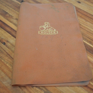 May include: A tan-colored Holden booklet with a gold emblem featuring a lion and the Holden name. The booklet is slightly worn and rests on a wooden surface with a diagonal pattern.