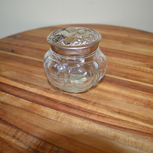 Op de afbeelding: Een heldere glazen pot met een zilveren deksel. De pot heeft een afgeronde vorm met een decoratief patroon. Het deksel is zilver met een bloemenmotief. De pot staat op een houten oppervlak.