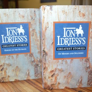 Puede incluir: Dos libros de tapa dura con cubiertas texturizadas de color marrón claro. Cada libro presenta un cuadrado azul con texto blanco y una silueta de una persona a caballo. El texto dice "Ion Idriess's Greatest Stories". Un libro dice "Heroes of the Outback", el otro dice "Of Miners and Soldiers".