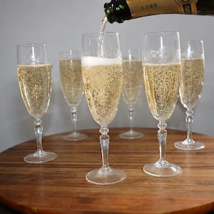 May include: Six clear glass champagne flutes arranged on a round wooden surface. A bottle labelled Brut is pouring sparkling golden liquid into the central glass, creating white foam. Each flute features a slender stem and a tall, narrow bowl.
