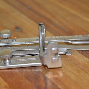 May include: A vintage metal edge binder tool with a ruler and adjustable knobs. The tool is made of silver-colored metal and is designed for binding edges. It is resting on a wooden surface.