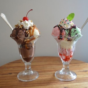 May include: Two glass sundae dishes filled with scoops of ice cream. The left dish contains chocolate and vanilla ice cream with chocolate syrup, caramel, whipped cream, nuts, and a cherry. The right dish has strawberry and mint ice cream.