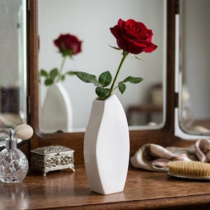 Puede incluir: Una rosa de color rojo intenso con hojas verdes colocada en un jarrón de cerámica blanca lisa. El jarrón tiene una forma asimétrica y estrecha, y se mantiene erguido. La rosa está en plena floración con pétalos visibles y un tallo largo.