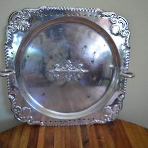 May include: A silver-colored, square serving tray with ornate, scrolled edges and two small handles. The tray has a slightly concave center and a decorative embossed design. The tray is sitting on a wooden surface.