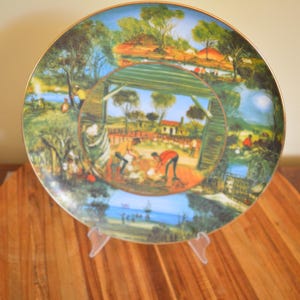 May include: Ceramic decorative plate with a gold rim, featuring a detailed landscape scene with figures. The plate has a central image framed by a circular border, depicting a rural setting with trees, buildings, and people. It is displayed on a clear plastic stand.