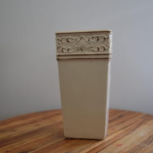 Puede incluir: Un jarrón de cerámica de color blanco roto, alto y de forma rectangular con una base estrecha. La sección superior presenta un patrón decorativo en relieve con volutas intrincadas y motivos florales que rodean todo el borde superior.