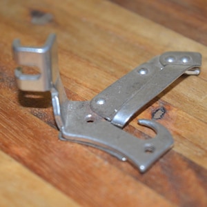 May include: A vintage silver can opener with a hinged design. The opener has a hook and a lever, designed for opening cans. The metal tool is shown on a wooden surface.