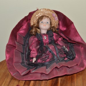 May include: A porcelain doll with brown hair and blue eyes wearing a straw hat with pink floral accents. The doll is dressed in a deep burgundy satin gown featuring black lace trim, a high collar with white pearl buttons, and a wide, ruffled skirt.