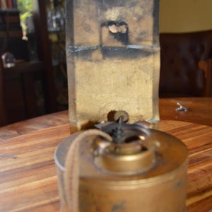 May include: An antique brass oil lamp with a cylindrical base and a rectangular back. The lamp has a wick and a small opening at the top. The back is gold with black accents and a hole near the top.