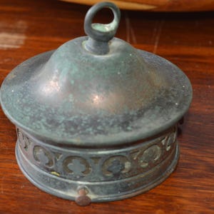 Może przedstawiać: Mały, zabytkowy metalowy pojemnik z pokrywką i uchwytem w kształcie pętli. Podstawa ma ozdobny, wzorzysty pas. Metal ma zwietrzałą, zielono-szarą patynę, wskazującą na wiek. Prawdopodobnie z brązu.