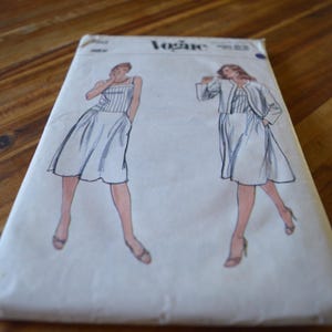 May include: A vintage Vogue sewing pattern. The envelope is white and features illustrations of a woman in a dress and jacket. The dress has a striped bodice and a flared skirt. The word "Vogue" is printed at the top of the envelope.