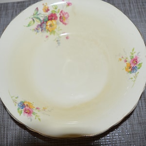 Op de afbeelding: Crèmekleurige, vintage serveerschaal met een geschulpte rand en gouden rand. De schaal is versierd met bloemclusters in roze, geel, blauw en paars. De schaal staat op een grijze ondergrond.