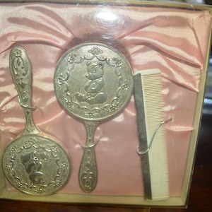 Puede incluir: Un juego de aseo para bebés vintage en una caja. El juego incluye un espejo de mano, un cepillo y un peine. Los artículos plateados presentan un diseño de oso de peluche. La caja está forrada con satén rosa.