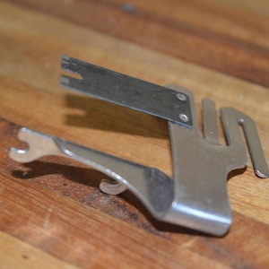 May include: A silver metal tool with a slotted rectangular top, a curved lever, and a forked end. The tool is designed for a specific purpose, possibly for opening or manipulating something. It rests on a wooden surface.