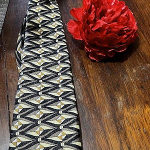 Può includere: Una cravatta fantasia con un design geometrico in nero, beige e oro, annodata in alto. Un vivace fiore rosso è appoggiato accanto alla cravatta su una superficie di legno. Il motivo della cravatta presenta forme a freccia ripetute.