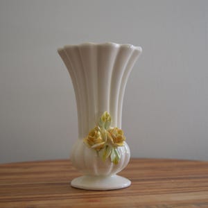 以下が含まれることがあります： クリーム色の陶器製花瓶で、縦に溝が入ったリブ状のデザインが特徴です。花瓶には広がった縁、丸みを帯びた台座があり、本体の前面には緑の葉がついた黄色いバラの立体的な装飾が施されています。