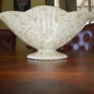 May include: A cream-coloured, mid-century modern ceramic vase with an unusual, asymmetrical shape. The vase has a speckled, web-like pattern in shades of brown and sits on a pedestal base. The vase is displayed on a dark wooden table.