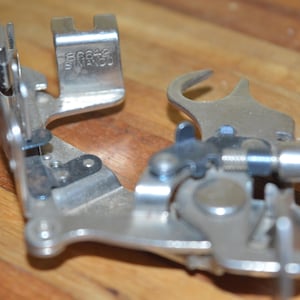 May include: A close-up of a silver sewing machine foot. The foot has multiple parts, including a lever, a screw, and a small metal plate. The foot is designed to attach to a sewing machine and guide fabric.