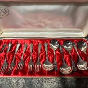 May include: A vintage silverware set in a cream-colored box with a red satin lining. The set includes forks, spoons, and serving spoons with ornate handles. The box is labeled "Superware Rodal".
